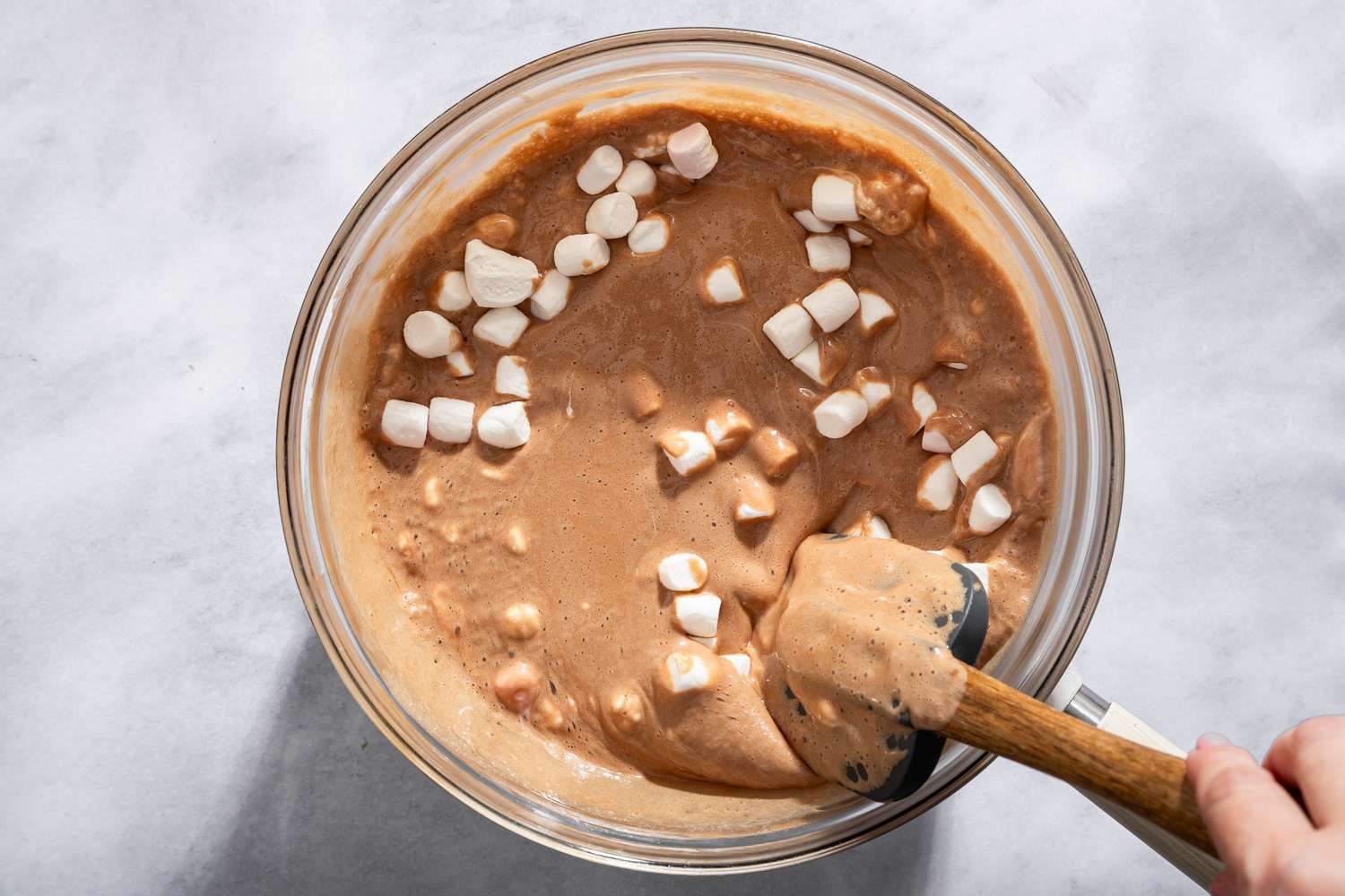 A glass bowl containing a chocolate mixture with marshmallows being stirred with a spoon