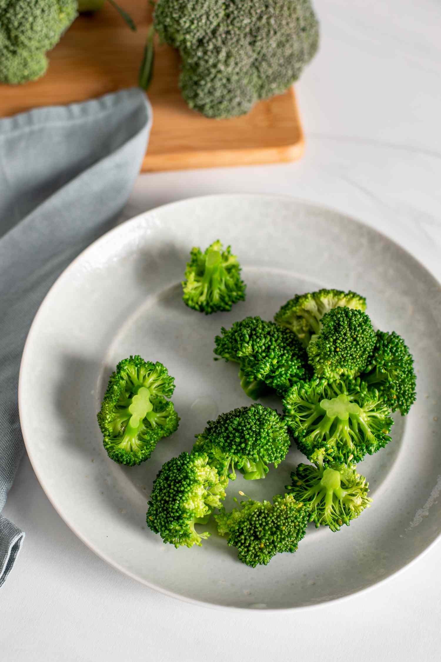 Blanched Broccoli on a Plate Surrounded by a Kitchen Towel and a Cutting Board with Heads of Broccoli for How to Blanch Broccoli