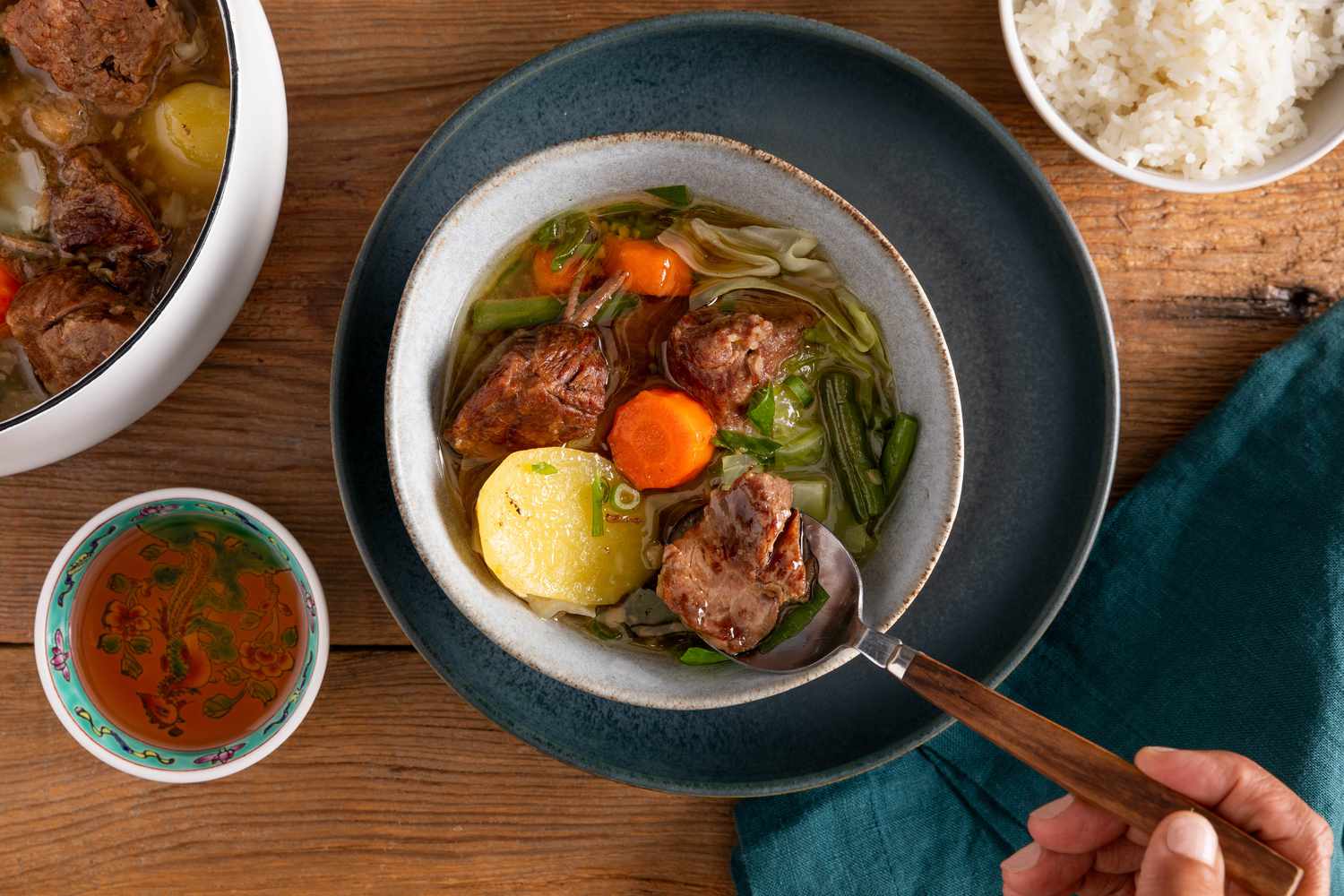 Serving of nilagang baka (Filipino beef stew) in a bowl, and in the surroundings, a Dutch oven with more soup, a small bowl of fish sauce, a bowl of rice, and a navy blue table napkin