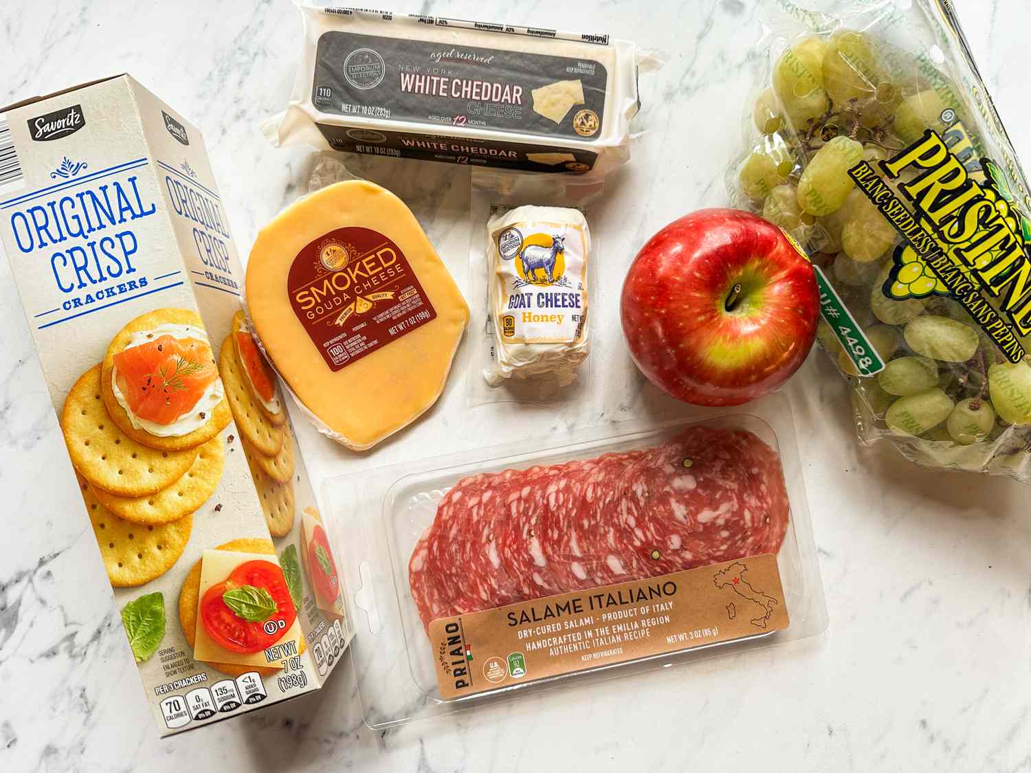 A selection of ingredients including crackers smoked gouda cheese white cheddar goat cheese salami an apple and grapes arranged on a marble surface