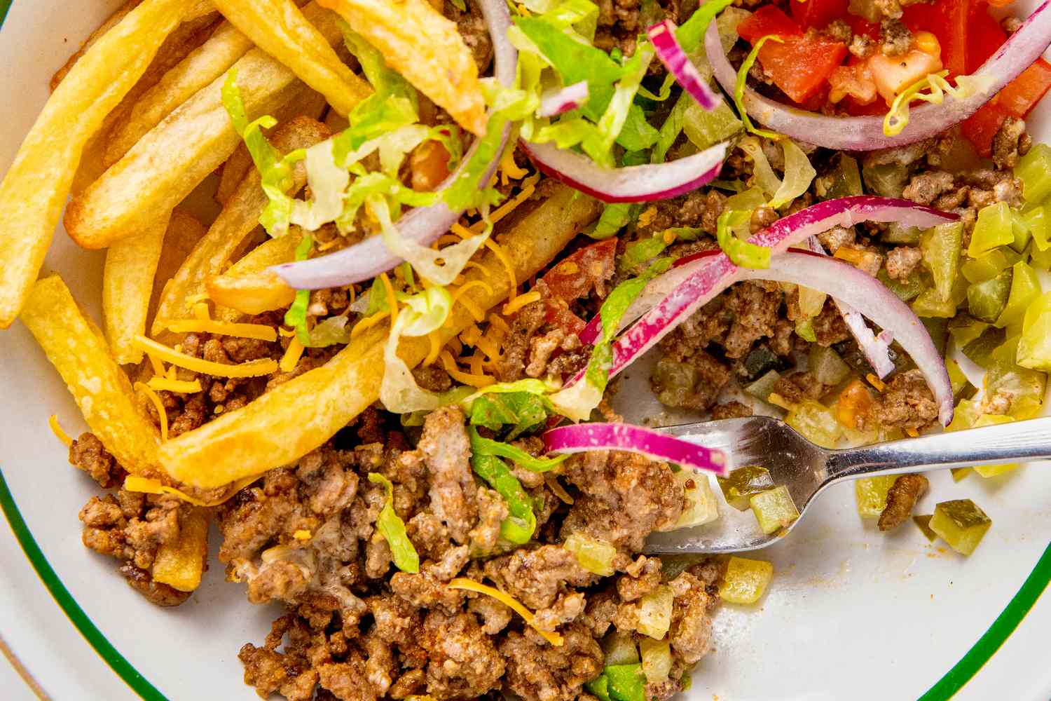 A burger bowl meal with ground beef lettuce onion tomato pickles and fries on a plate with a fork