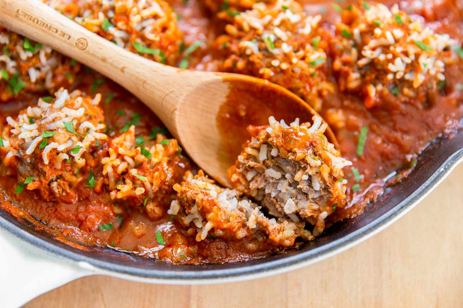 Side view of a cast iron skillet of meatballs with expanded rice on the surface and sauce on a wooden cutting board with half of a meatball being scooped out by a wooden spoon 