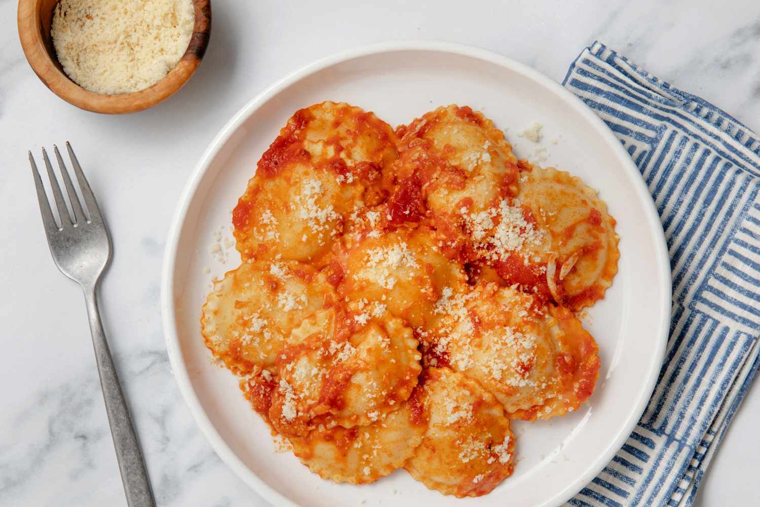 Dish of ravioli with red sauce and grated cheese on a white plate, fork and cloth napkin beside it