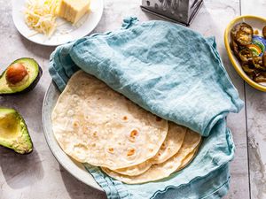 Homemade flour tortillas on a plate in-between a teal linen. A small bowl of pickled jalepanos, a box shredder, shredded sheese and a halved avocado are around the plate.