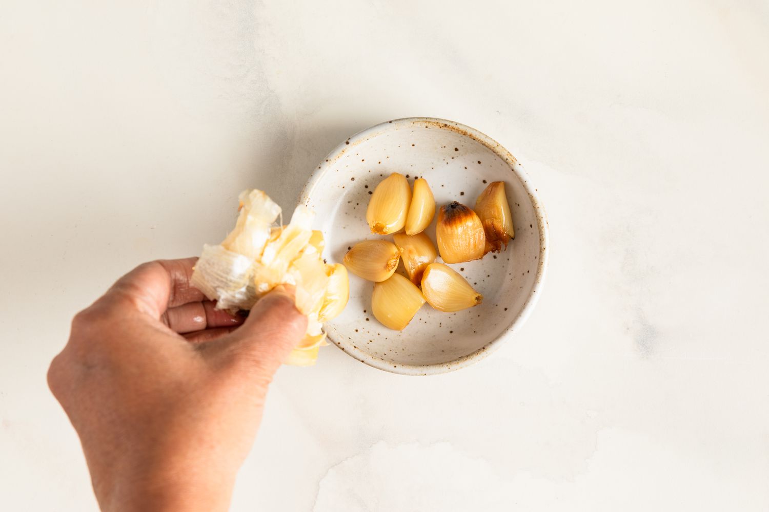 Roasted garlic cloves in a bowl being squeezed out of their peels by a hand