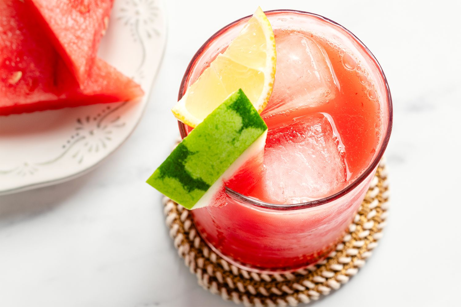 A glass of watermelon lemonade with ice garnished with a lemon and watermelon slice next to a plate of watermelon wedges