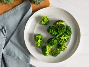 Blanched Broccoli on a Plate Surrounded by a Kitchen Towel and a Cutting Board with Heads of Broccoli for How to Blanch Broccoli