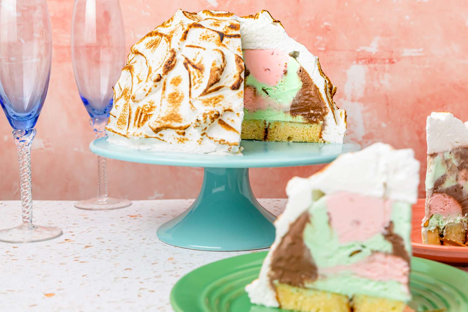 Baked Alaska with Slices Missing on a Cake Stand, and in the Surroundings, Two Plates with Slices of Baked Alaska and Champagne Flutes