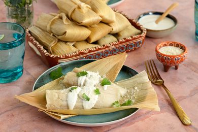A Plate of Chicken Tamales with Chile Verde Topped with Crema, Cotija Cheese, and Some Cilantro at a Table Setting with a Platter of More Tamales, Utensils, a Small Bowl of Cotija Cheese, a Small Bowl of Crema, a Glass of Water, and a Small Vase