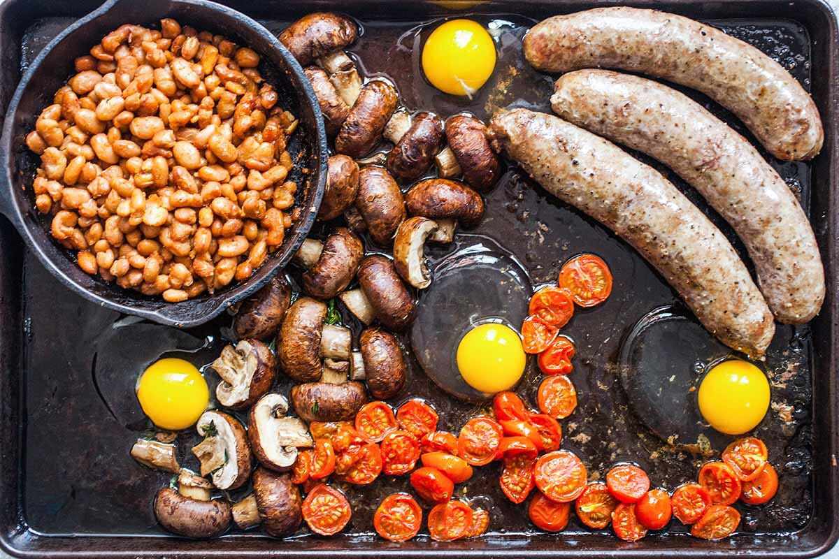 Eggs cracked on a sheet pan with sausages, tomatoes, potatoes, and a pot of beans