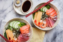 Two Chirashi Bowls with Salmon, Tuna, Shrimp, Tamagoyaki, and Sliced Cucumbers Next to a Small Saucer of Wasabi and Another Small Saucer of Soy Sauce