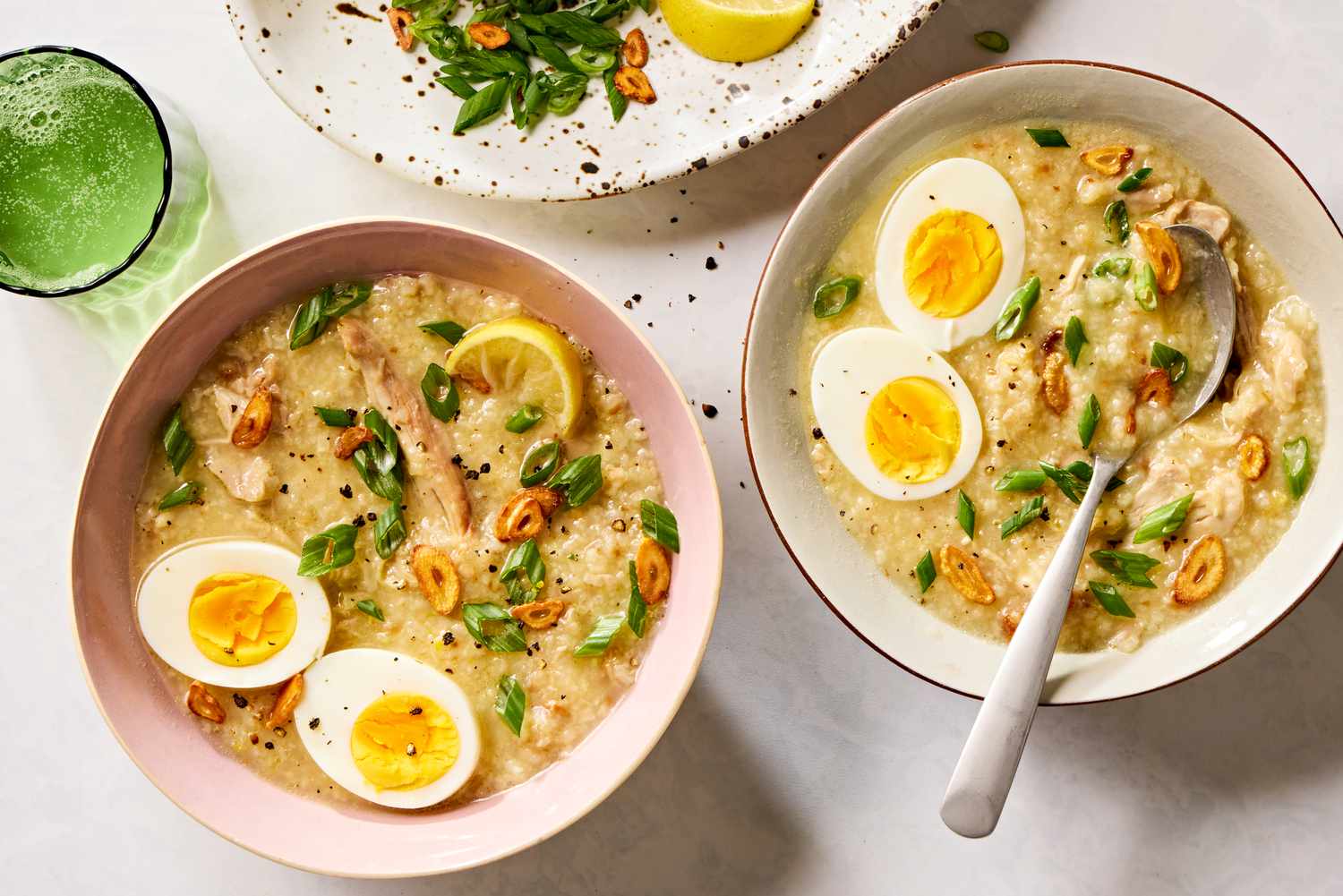 Overhead view of two bowls of Arroz Caldo with Chicken recipe, one with a spoon, all next to a green glass and plate of lemon slices and scallions 