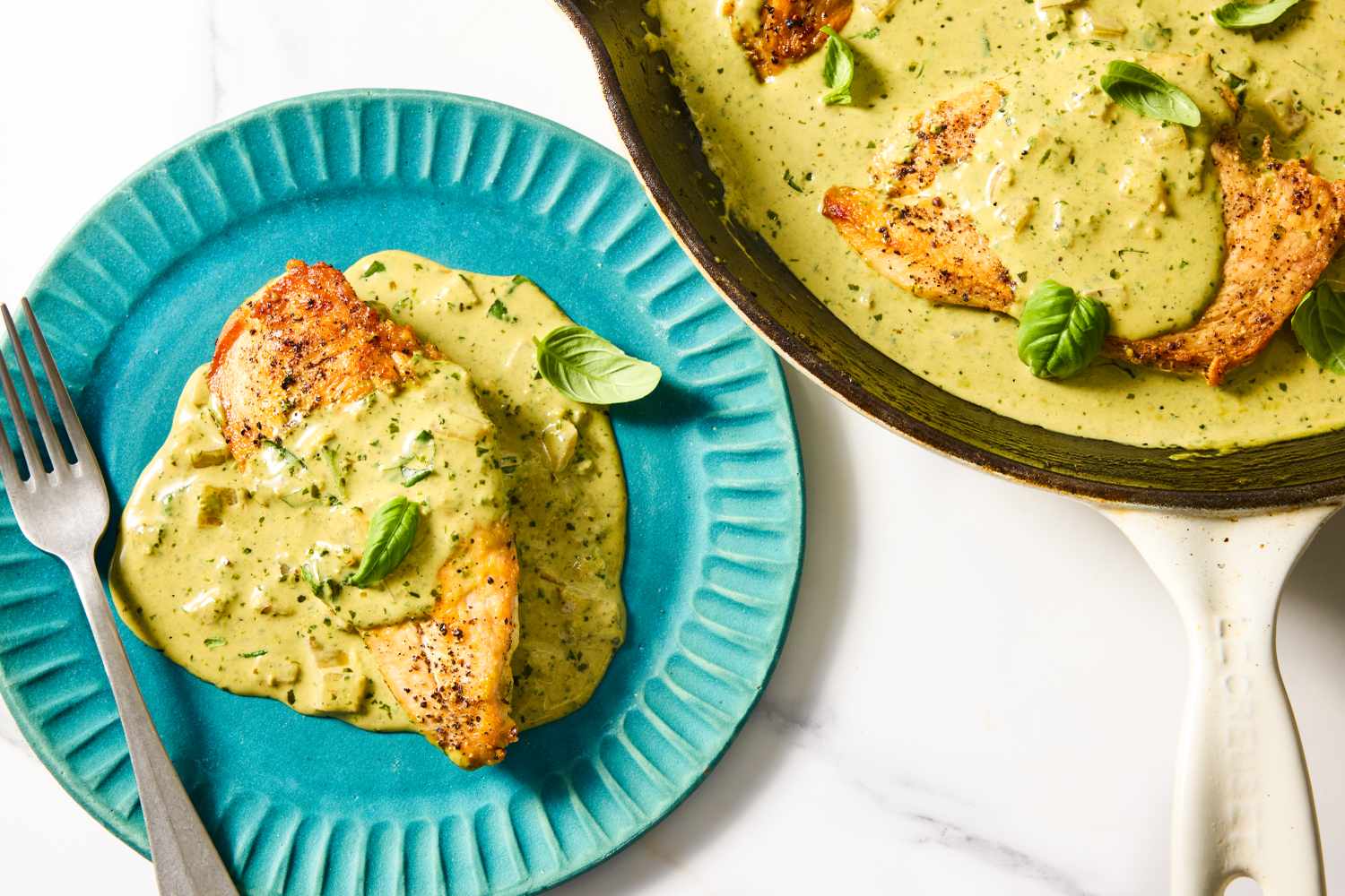 A plate with a chicken breast covered in pesto sauce next to a skillet containing more chicken in sauce garnished with basil leaves