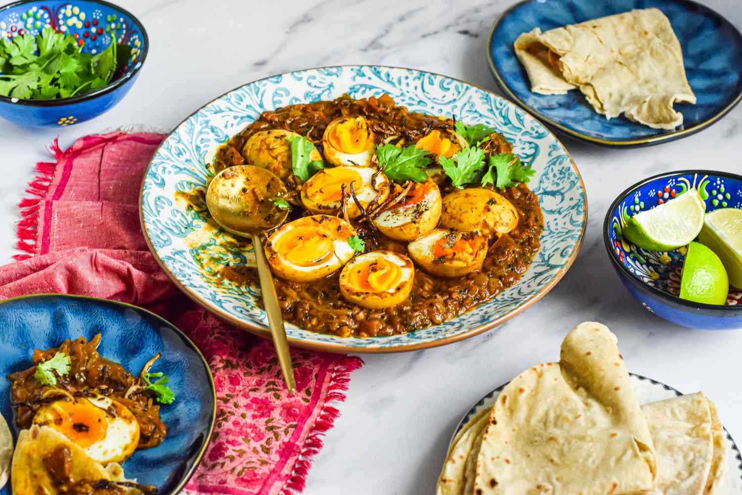 Plate of Egg Masala Curry with a Spoon on a Kitchen Linen, Surrounded by a Plate with a Serving of Curry and Roti, Another Plate with More Roti, a Bowl with Lime Wedges, and a Bowl with Cilantro