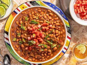 borracho beans (frijoles beans) in a bowl at a table setting with a bowl of diced tomatoes, small plate with lime wedges, a glass of beer, and utensils