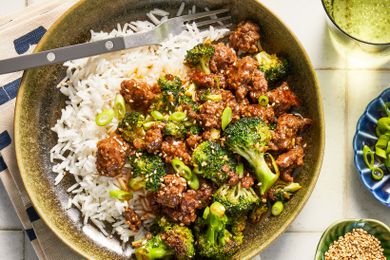 Ground beef and broccoli stir fry over rice in a bowl at a table setting with a glass of water, a bowl of sliced scallions, and a bowl of sesame seeds