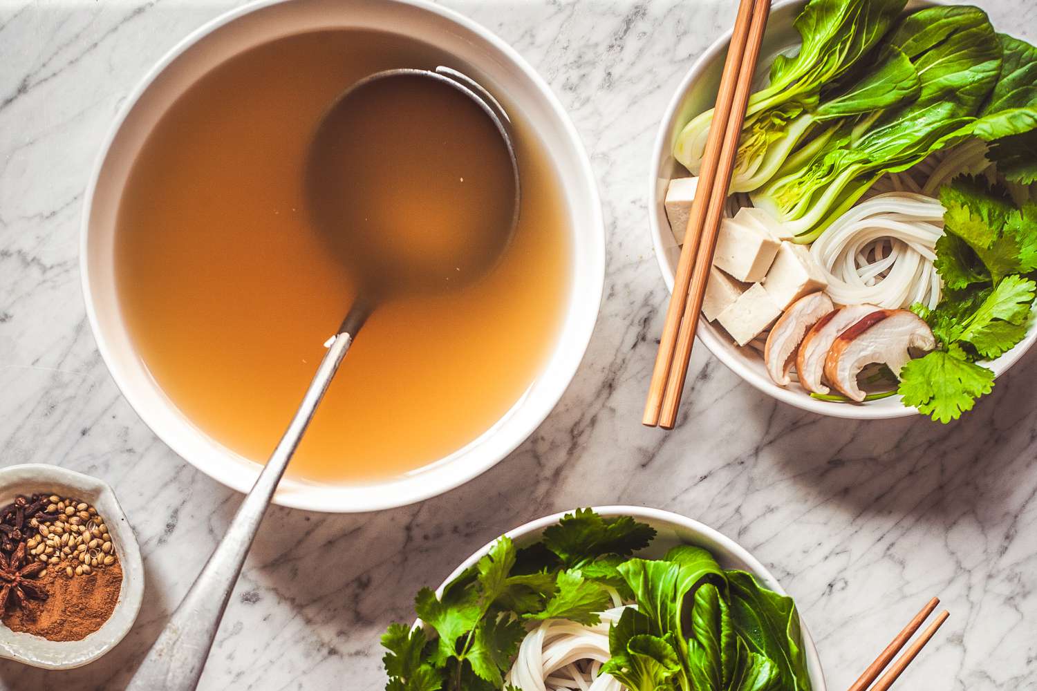 Overhead view of a white bowl of broth with a ladle in it next to bowls of greens, noodles, mushrooms, tofu with chopsticks and a bowl of spices