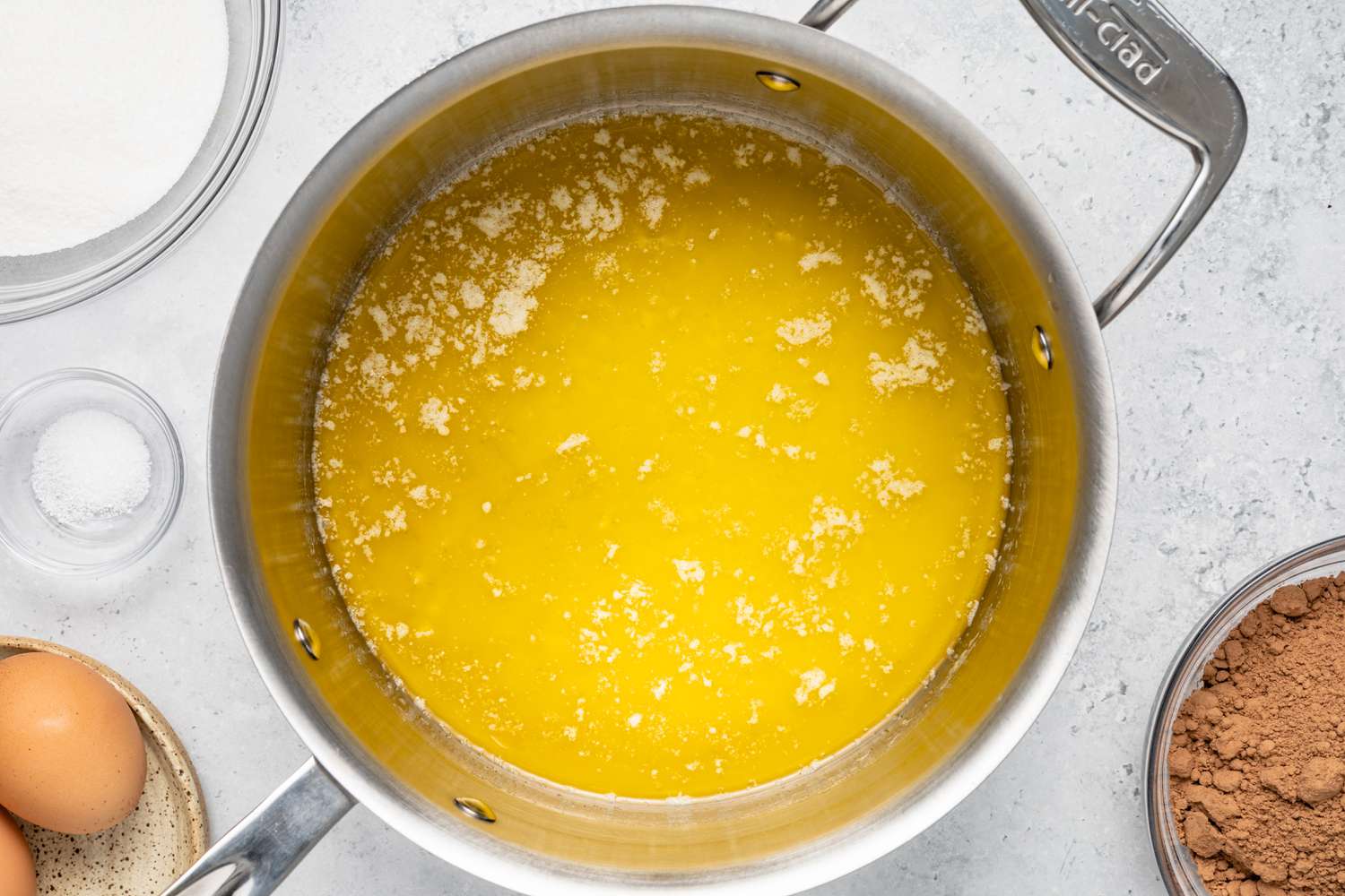 Pot of Melted Butter Surrounded by a Bowl of Cocoa Powder, a Small Bowl of Salt, a Saucer with Two Eggs, and a Bowl of Sugar for Brownie Banana Bread Recipe