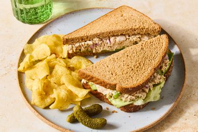 Small plate with a tuna salad sandwich, chips and small pickles on a tan countertop