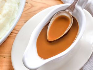 Overhead view of a gravy boat of gravy and a spoon full of gravy next to a plate of mashed potatoes and a white napkin all on a wooden tabletop