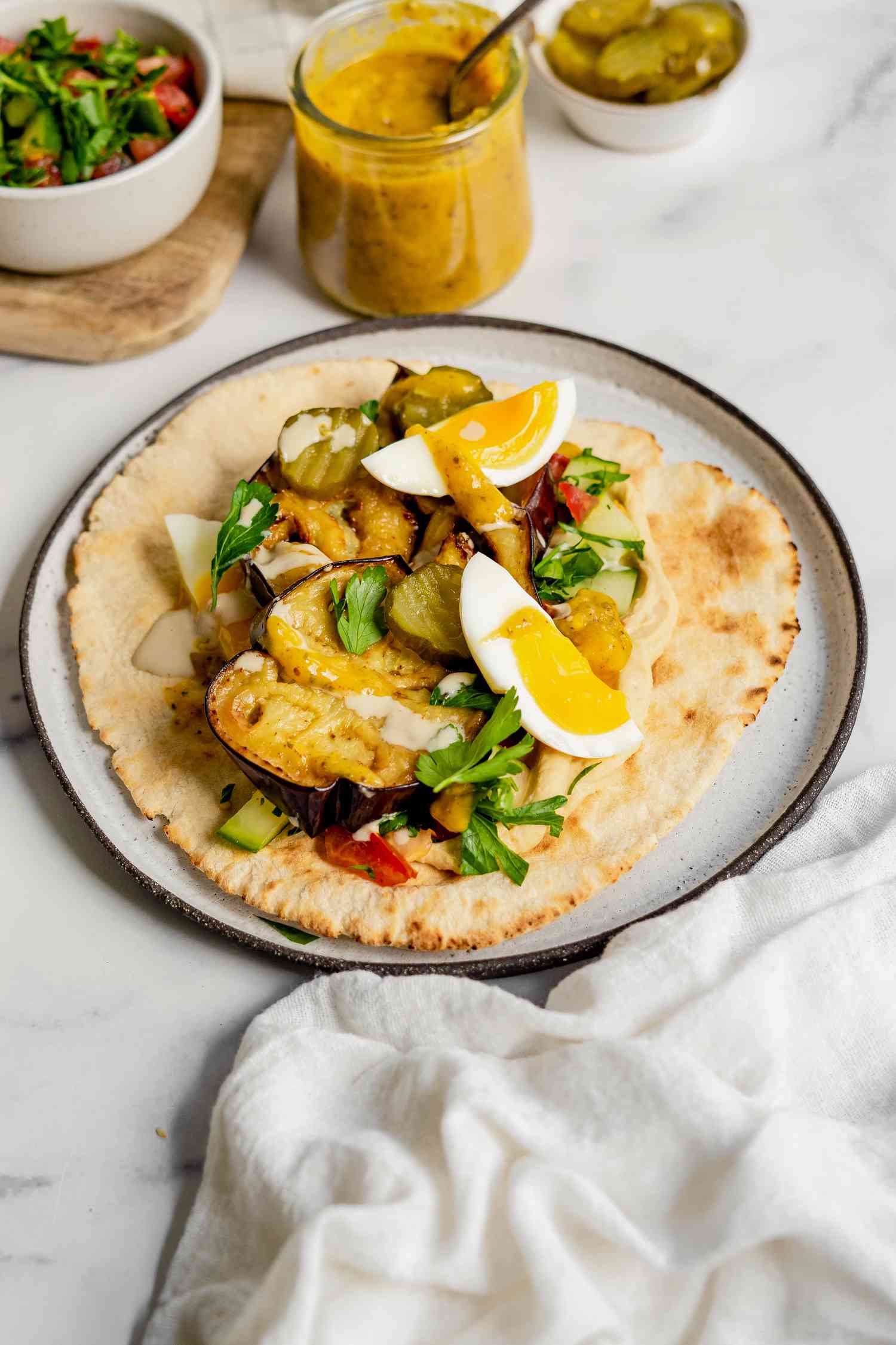 Sabich on a Plate Next to a Bowl of Israeli Salad and a Jar of Tahini Sauce