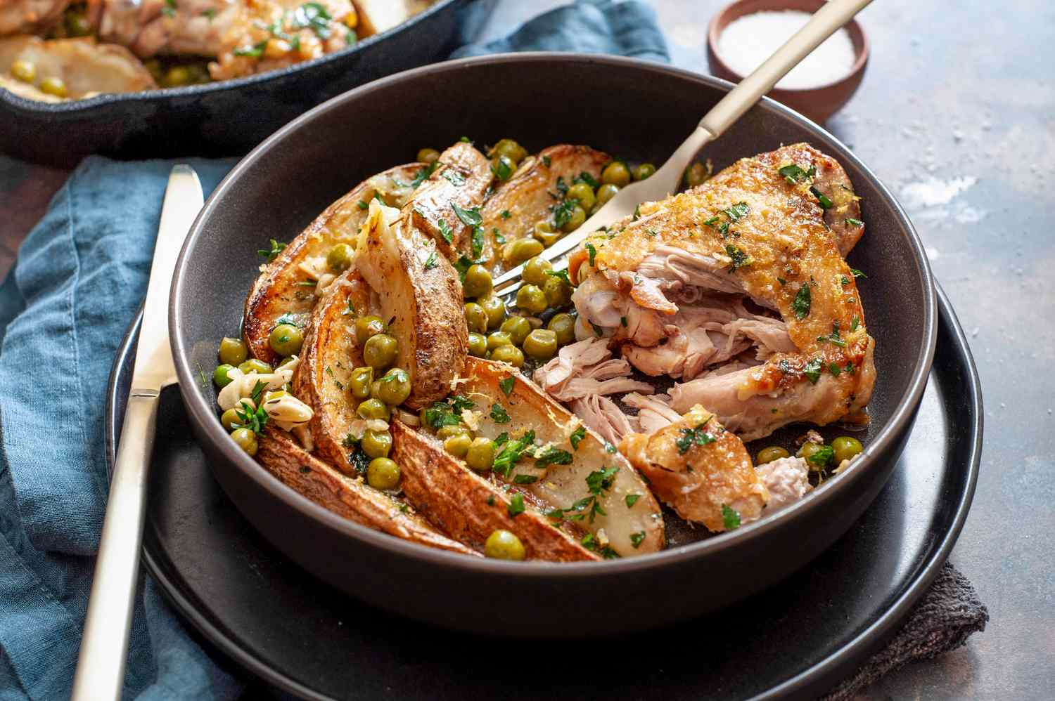 Chicken Vesuvio in a Bowl on a Plate Next to a Cast Iron Skillet with More Chicken Vesuvio and a Small Bowl of Salt, All Sitting on a Table Napkin