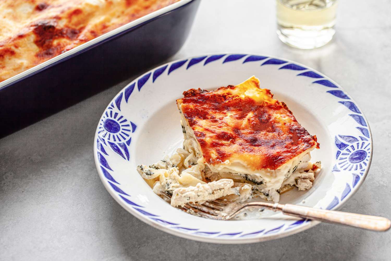 Slice of Chicken Lasagna in a Bowl with a Bite on a Fork, and in the Background, a Casserole Dish with More and a Glass of Water