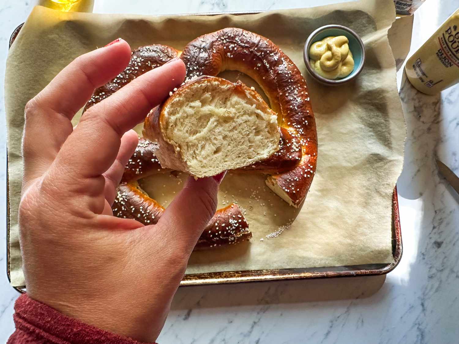 A hand holding a piece of a pretzel, tray with pretzels and mustard dip in the background