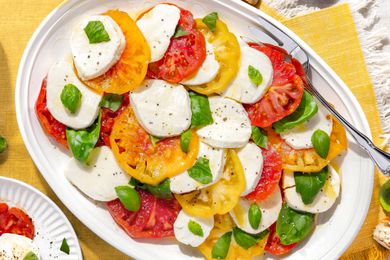Platter of Caprese Salad With Tomatoes, Basil, and Mozzarella Next to a Plate With a Serving, All on a Yellow Placemat