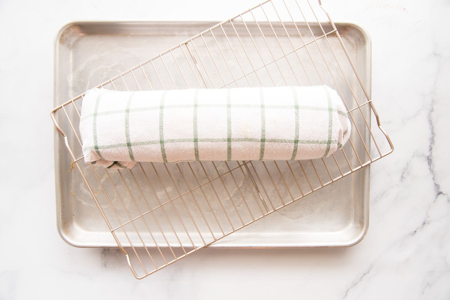A baking sheet with a cooling rack set at an angle and a swiss roll cake wrapped in a tea towel on top.