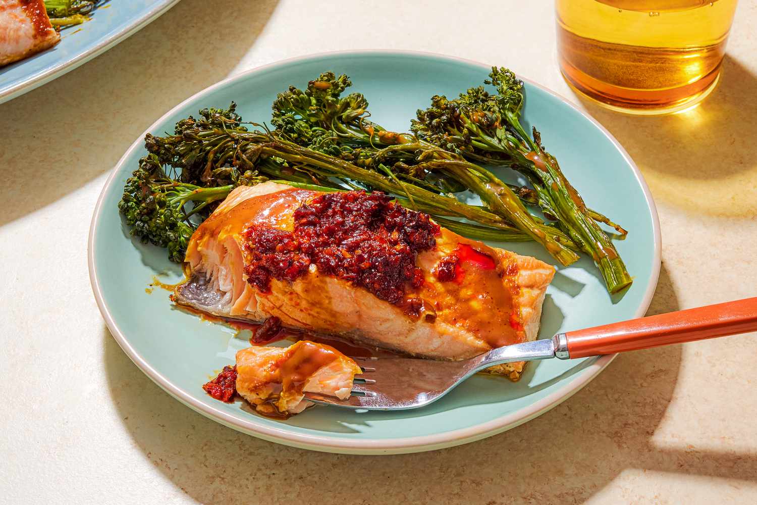 Angled view of a light blue plate of a cooked salmon filet topped with chili crisp and stems of broccolini with a fork holding a bite of salmon