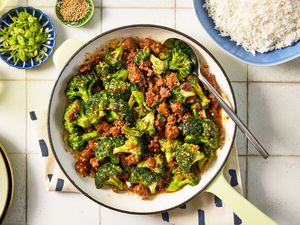 Bowl of ground beef and broccoli stir fry at a table setting with a bowl of rice, bowl of sesame seeds and a glass of water