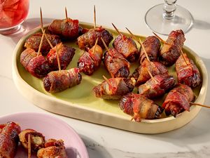 A tray of baconwrapped dates served with toothpicks accompanied by a glass of red drink in the background