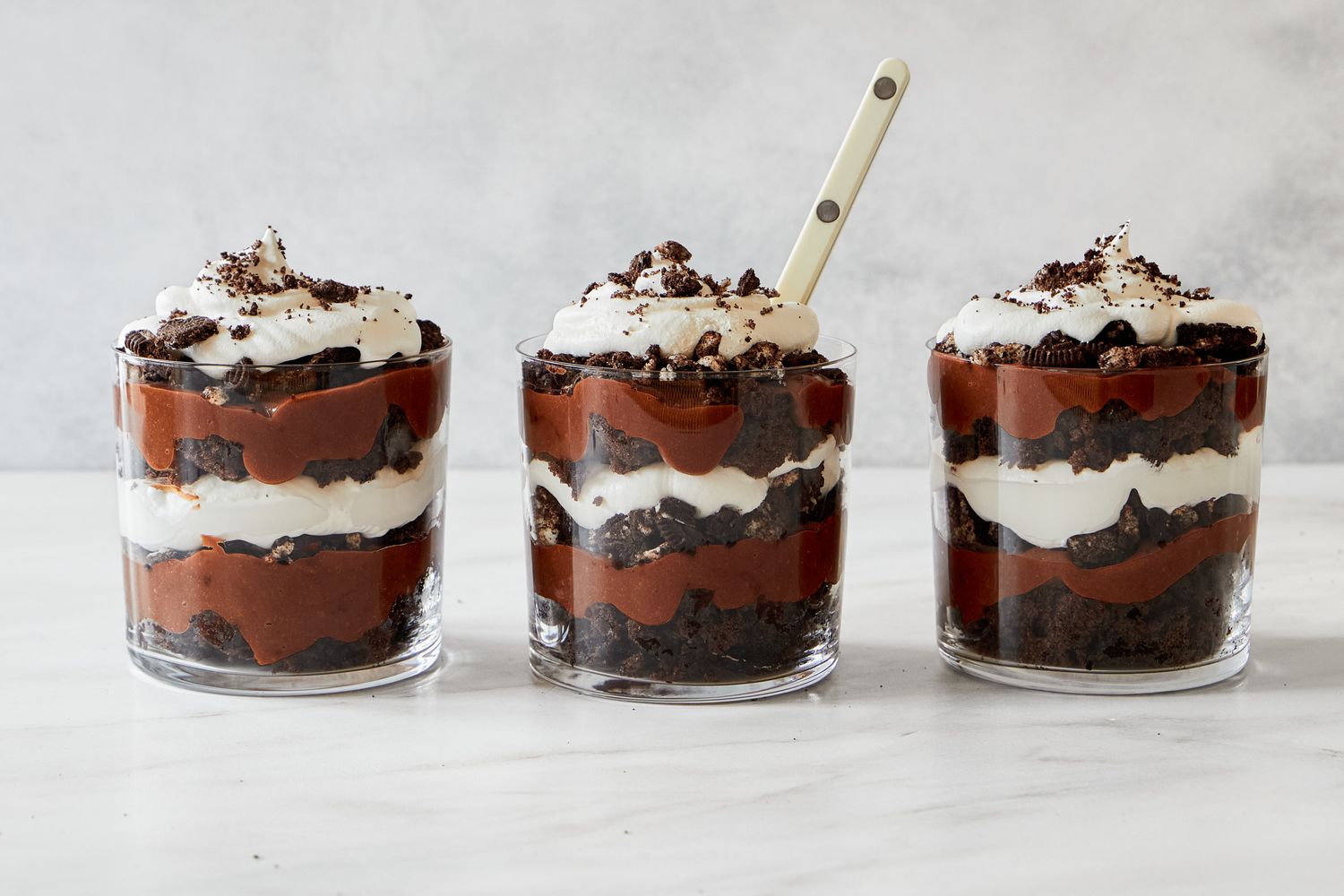 Three layered chocolate parfaits topped with whipped cream and crumbled toppings placed in glass containers on a white surface