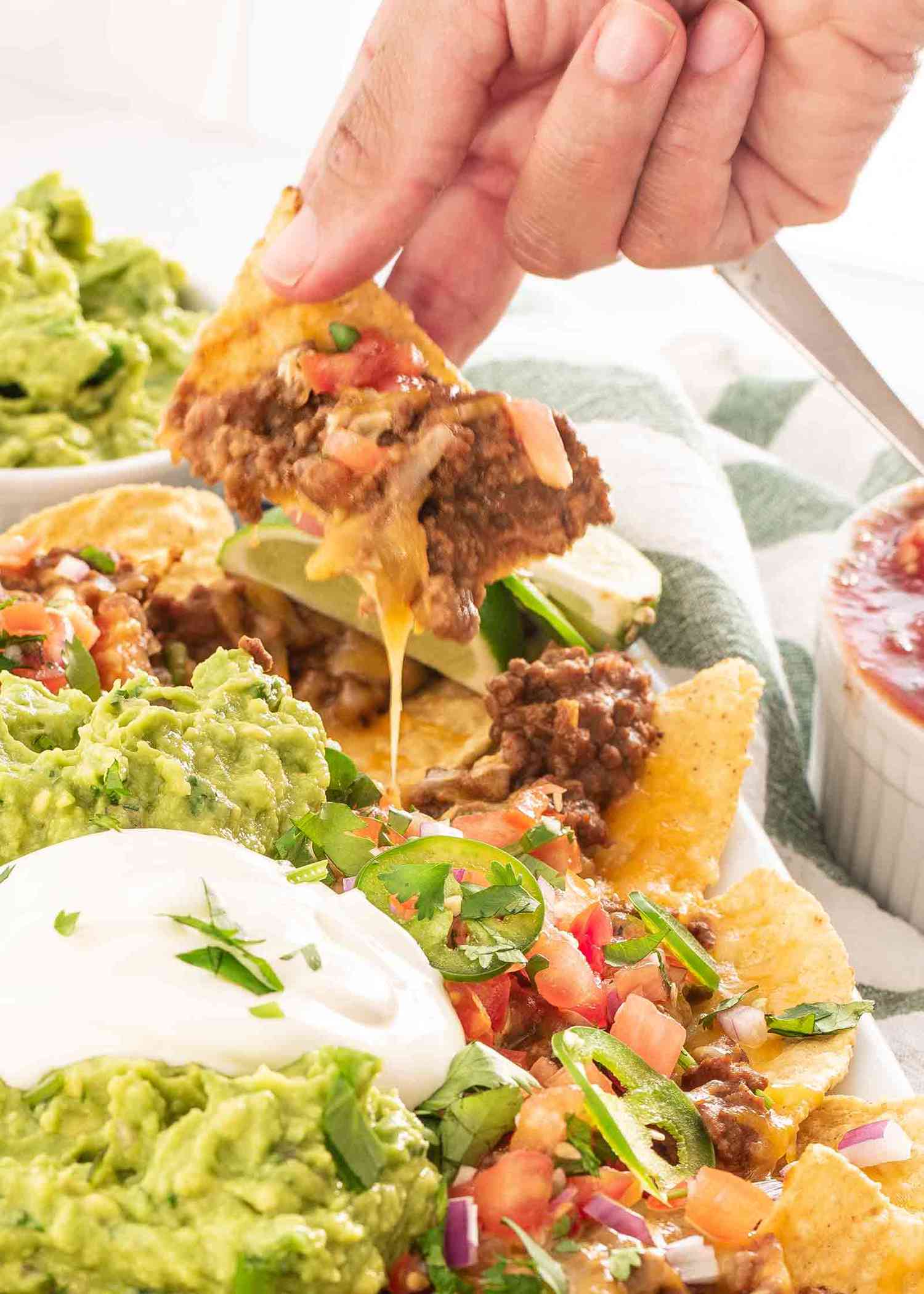 A hand taking a loaded nacho chip from a plate of nachos covered in guacamole and sour cream 