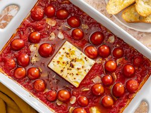 Overhead view of a white baking dish with garlic, tomatoes, cannellini beans and a block of feta covered in olive oil and red pepper flakes next to a plate of buttered french bread slices and a napkin