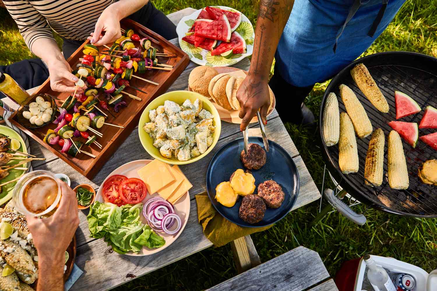 Outdoor meal with skewers, burgers, and grilled items on a picnic table near a grill