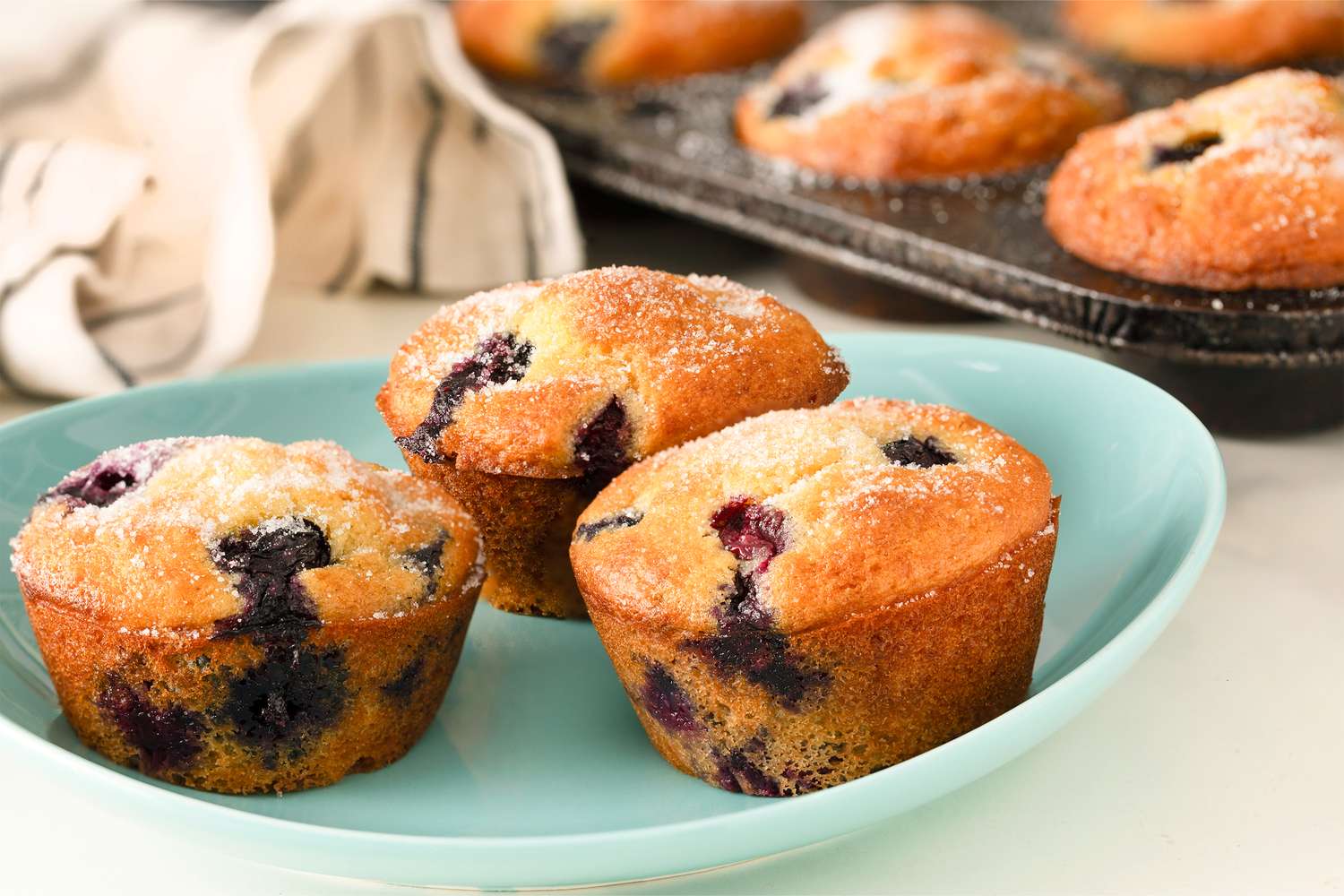 3 blueberry muffins on a small plate, muffin tin with muffins in the background