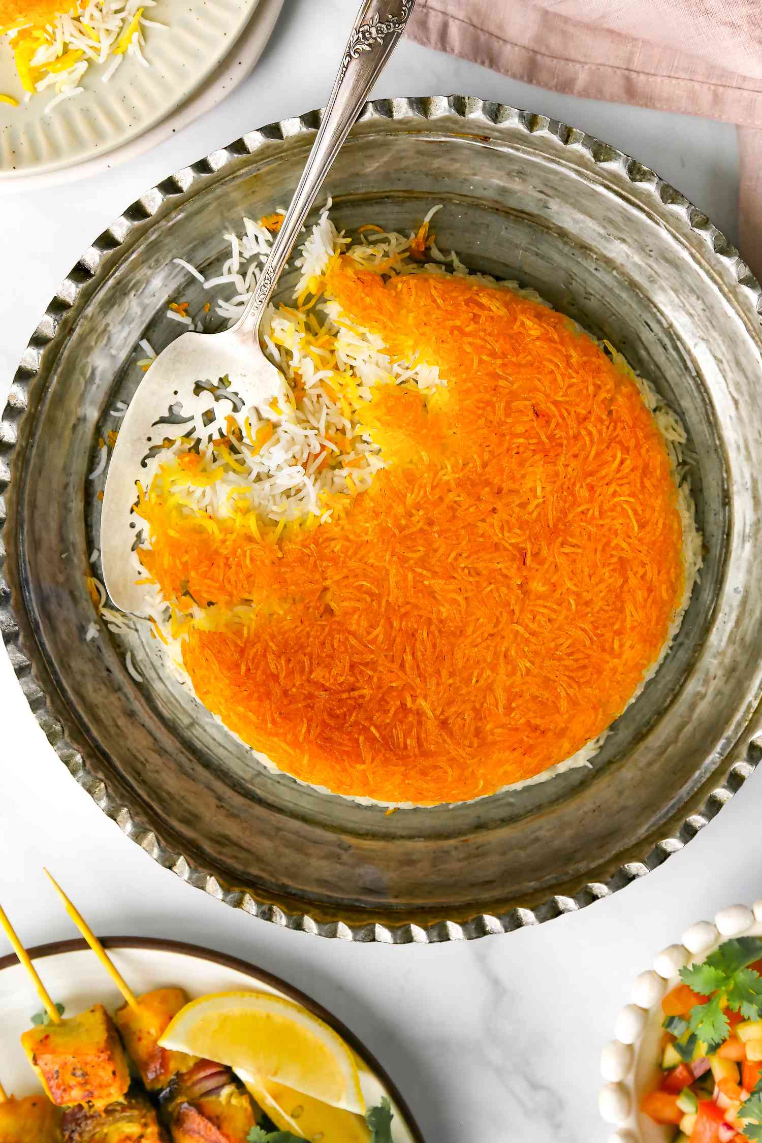 Serving Resting on the Side of Tahdig on an Ornate Silver Platter after Serving It, and Surrounding the Platter, Two Plates of Food (One with a Salad and Another with Kebabs)