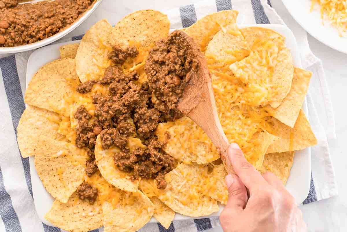 Refried beans and ground beef mixture being spooned on to cheese covered tortilla chips