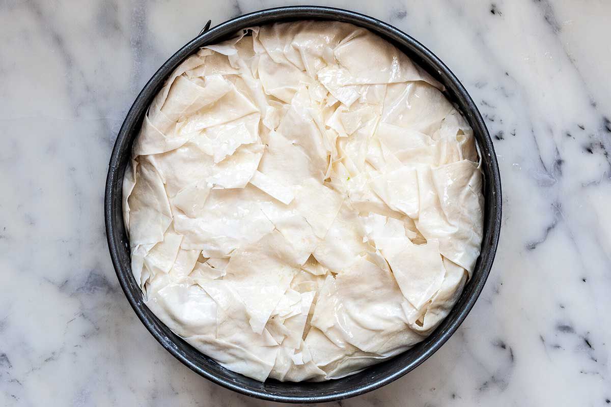 A cake pan is on a marble countertop. Unbaked phyllo dough is stacked and messily folded around the filling.