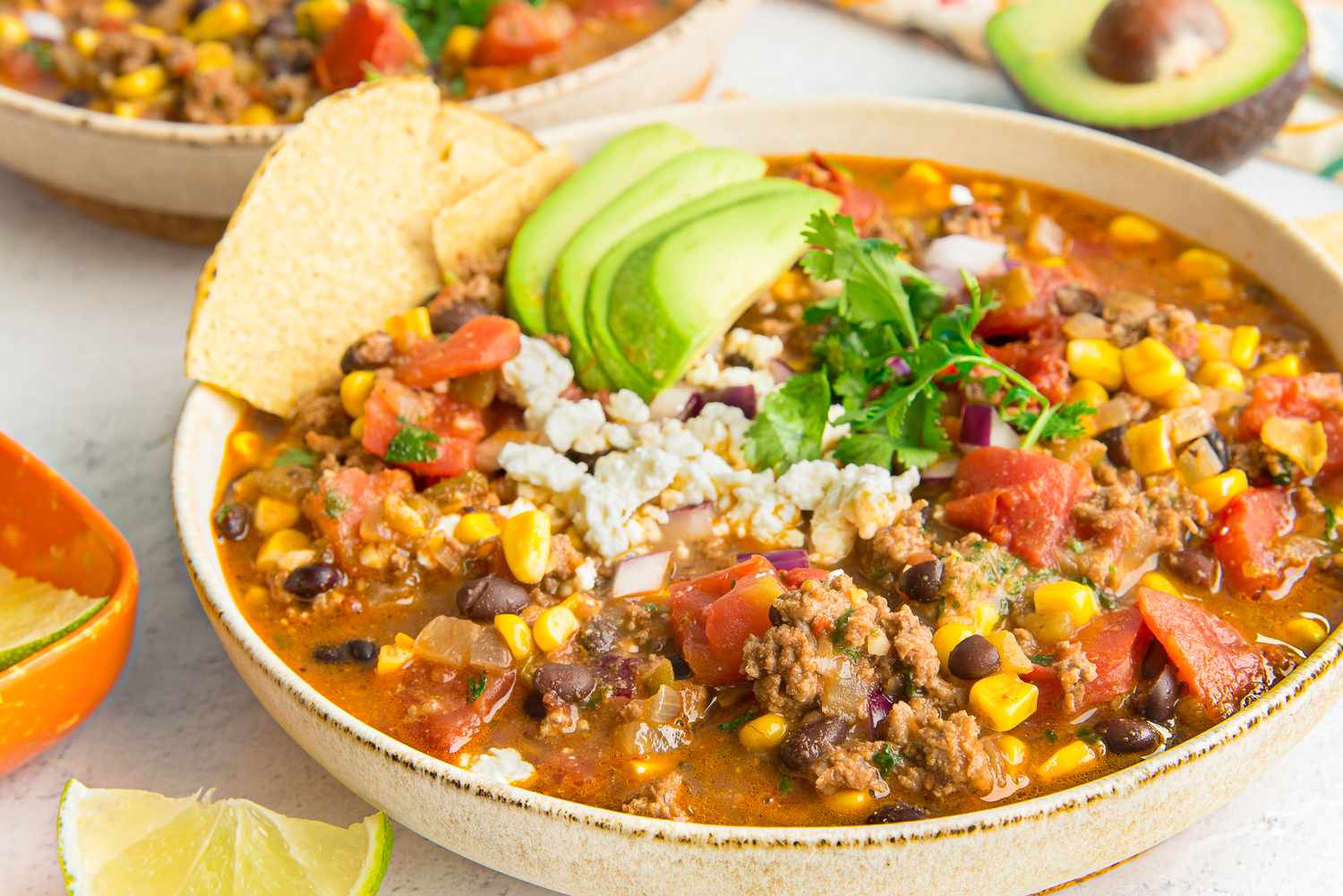 Bowl of Stovetop Taco Soup Topped with Avocado Slices, Cotija Cheese, and Cilantro and Surrounded by Bowls of Toppings