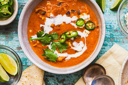 Creamy Chicken Enchilada Soup Topped with Jalapenos, Cilantro, and Crema in a Bowl at a Table Setting with Two Spoons on a Table Napkin, Bowls of Toppings (Jalapenos and Lime Wedges), and Tortillas