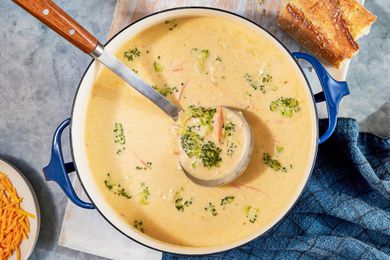Pot of copycat Panera broccoli cheddar soup with a ladle raising some of it, and in the surroundings, a bowl of shredded cheese, a piece of baguette, and a blue kitchen towel