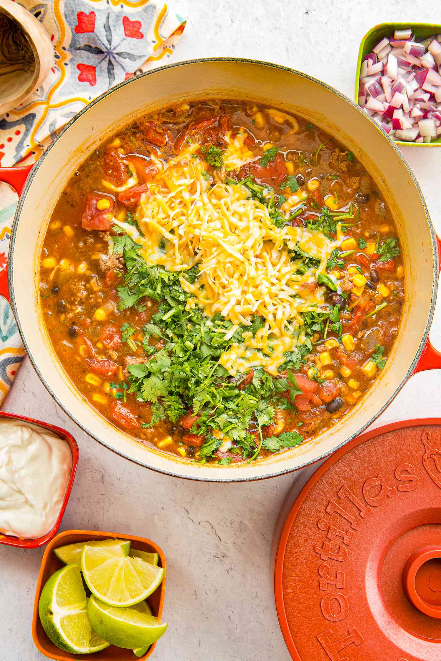 Stovetop Taco Soup in a Dutch Oven Topped with Cheese and Cilantro and Surrounded by Bowls of Sour Cream, Diced Red Onions, and Lime Wedges