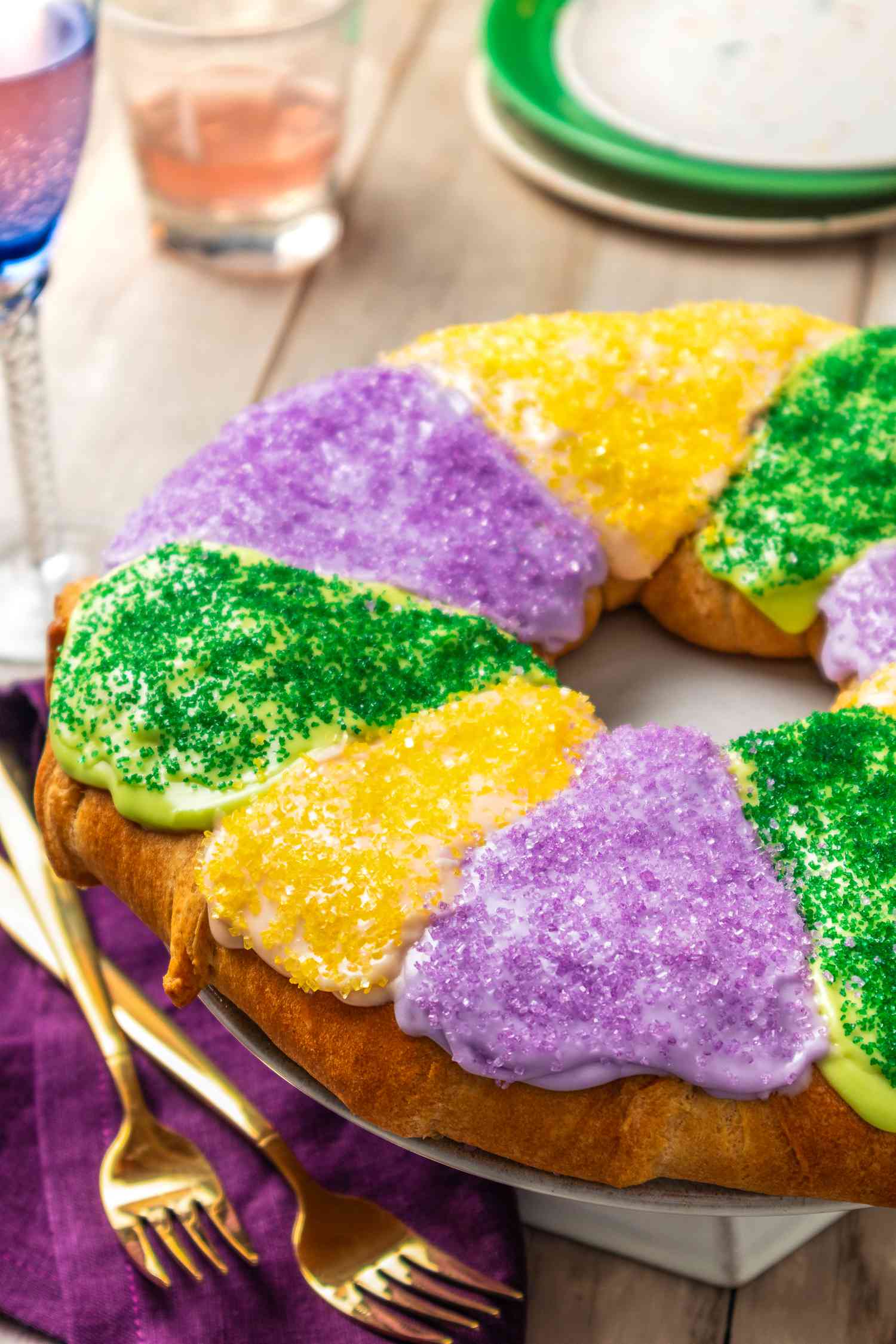 King cake (all decorated) on a cake stand at a table setting with utensils, a purple napkin, wine glasses, and a stack of plates