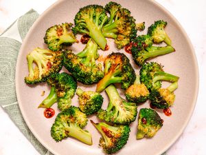 Overhead view of a pink plate of chili crisp coated broccoli florets resting on a cloth napkin and marble countertop