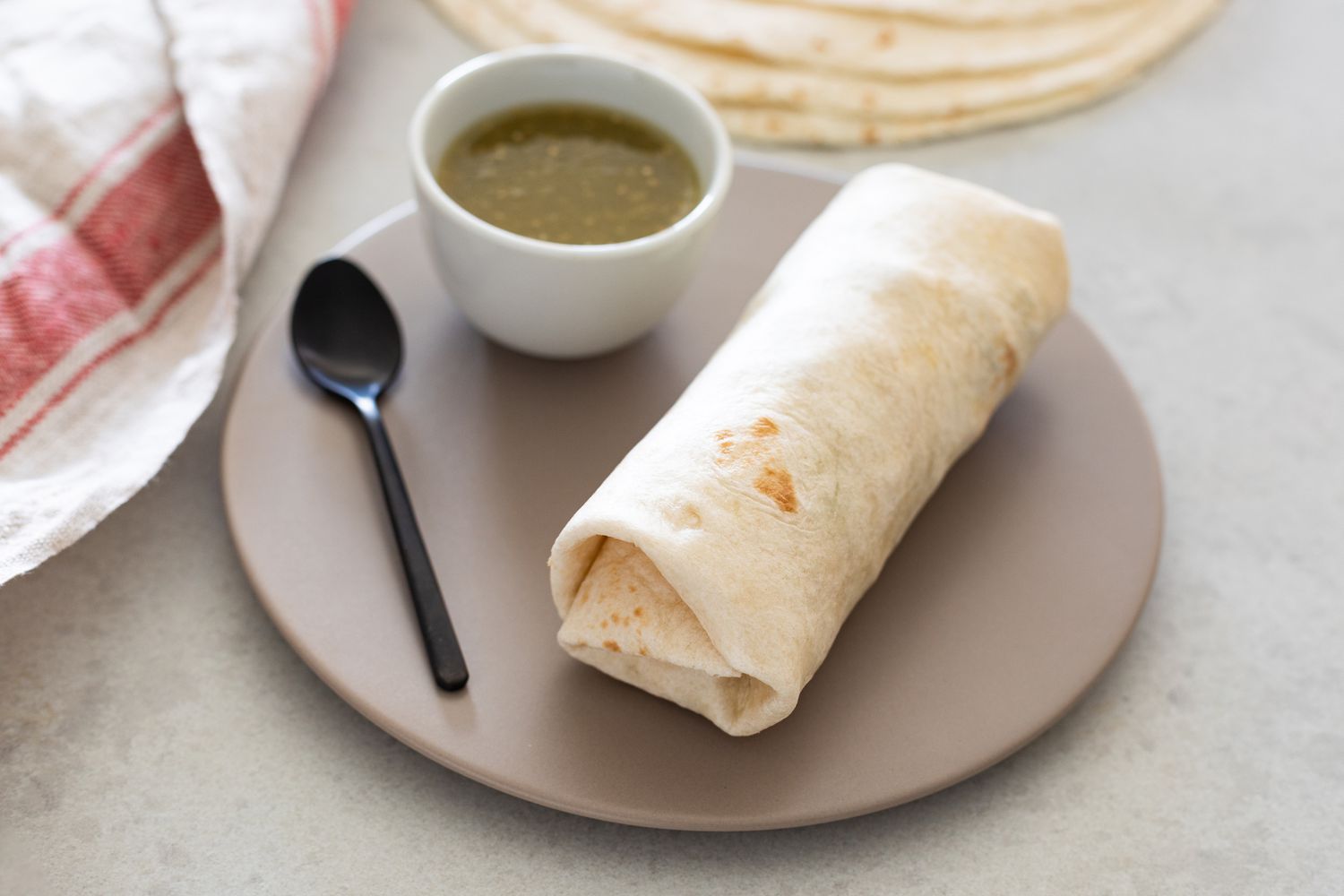 Nicely Folded Burrito on a Plate with a Small Bowl of Salsa and in the Background, a Stack of Tortillas