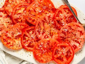Plated marinated tomato slices topped with seasoning and shredded cheese served on a white dish with a fork
