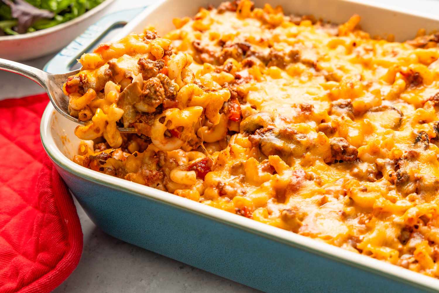 Spoonful of Johnny Marzetti Casserole lifted from the casserole dish, and in the surroundings, a red pot holder and a bowl of mixed greens salad
